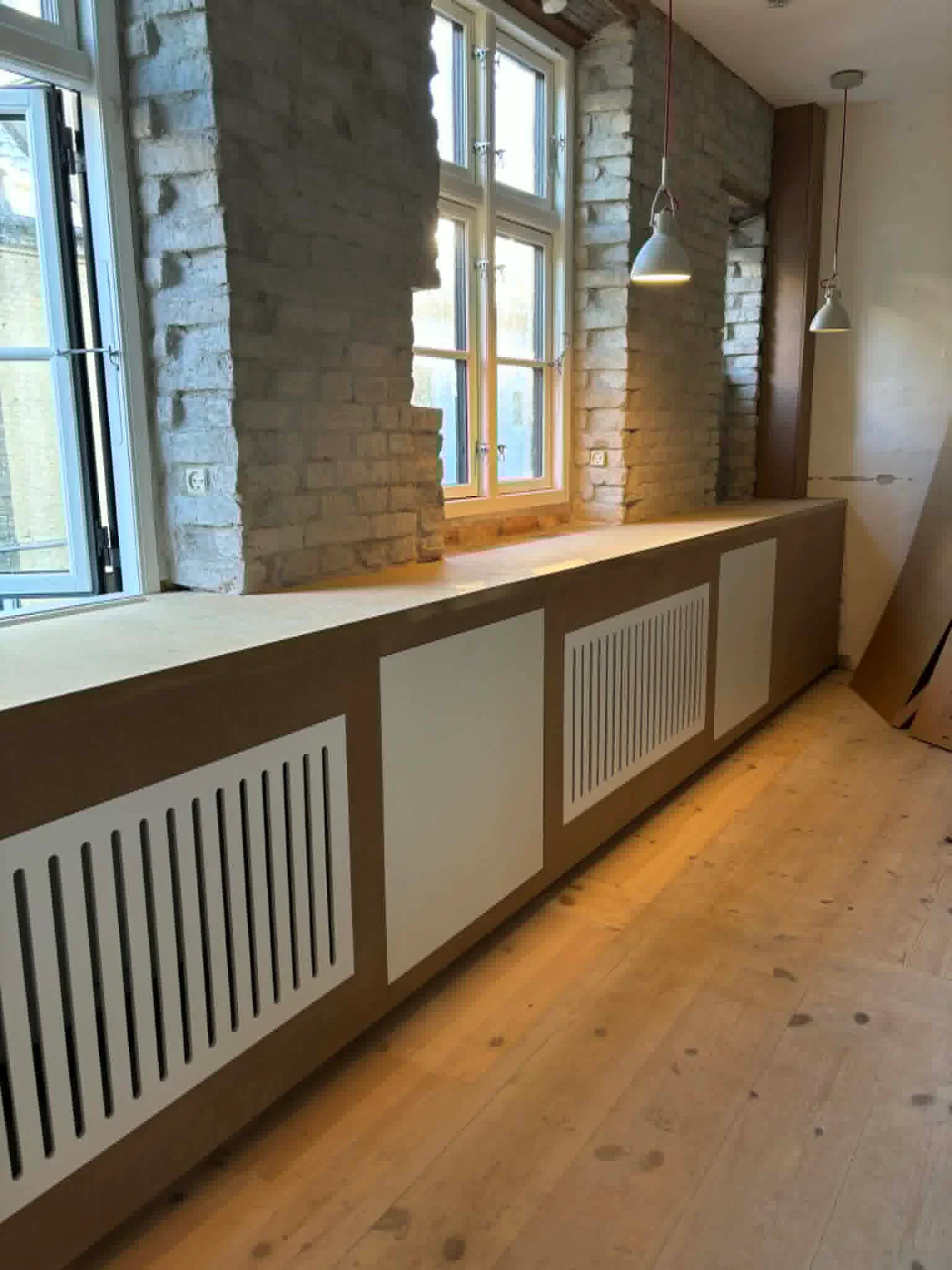 Håndlavet radiatorafskærm i træ under store vinduer i et indendørs rum.