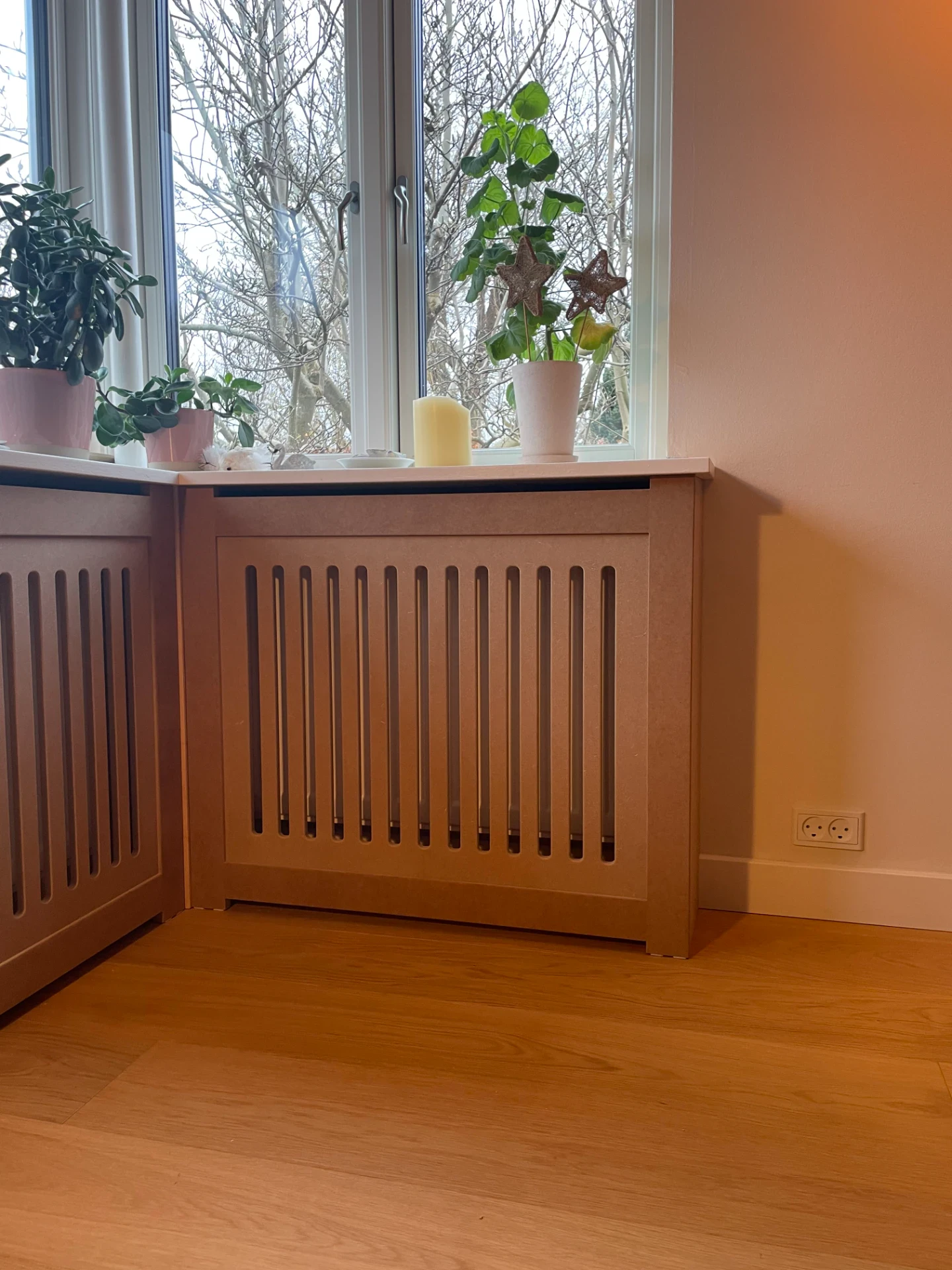 Håndlavet træradiatoromramning under vindue med planter og lys.